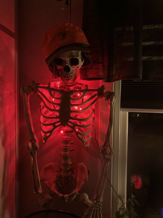 Ett skelett med bygghjälm i röd belysning framför ett fönster.