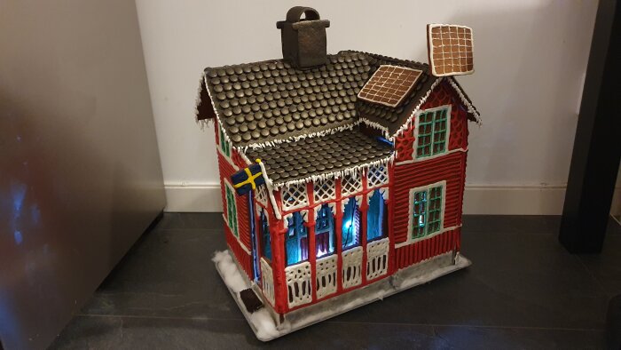 Ett pepparkakshus med detaljer, svensk flagga, snö, belysning, tak med kakel, skorsten, utsmyckade fönster och dörr.