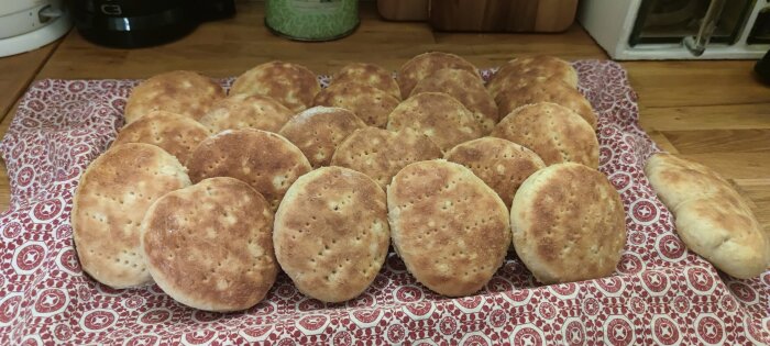 Nybakade, runda bröd på mönstrad kökshandduk, kanske hembakta bullar eller bröd, på köksbänk.