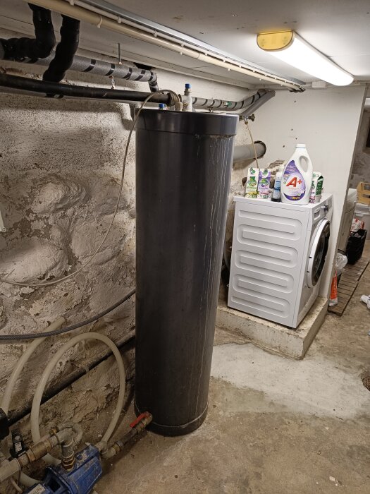 Tvättstuga med tvättmaskin, högt placerad vattenberedare, rengöringsmedel och betongväggar. Rör och avlopp synliga.