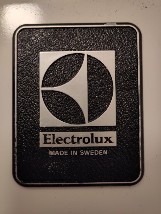 Elektrolux logotyp plakett, svart bakgrund, silvertext, ”Made in Sweden”, slitna kanter, reflektioner, troligen hushållsapparat.