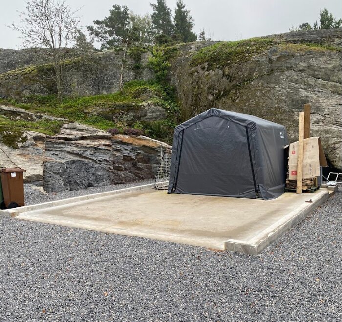 Betongplatta med uppsatt tältkonstruktion och förpackningsmaterial på grusig mark, inramad av klippvägg och grönska.