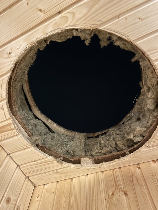 Rund hålighet i ett träpaneltak, exponerad isolering, mörker syns genom öppningen, potentiell renoveringsplats.