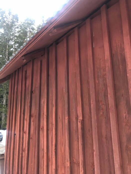 Rödmålad trävägg med stående paneler, hörn av hus, takkant synlig, grönska i bakgrunden.