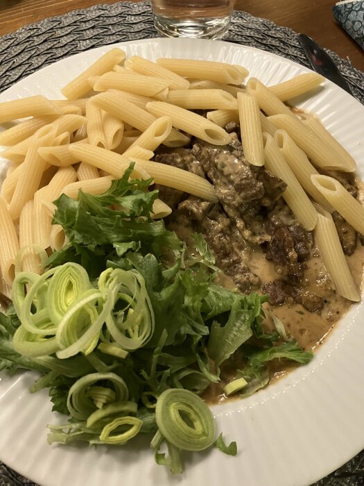 Pasta med sås, grönsallad och purjolök på ett vitt fat, ett glas vatten, en sked.