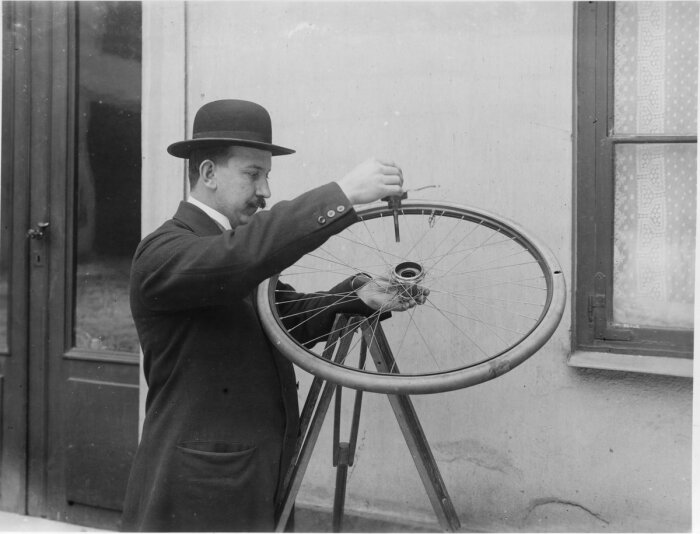 Man i kostym och hatt reparerar cykelhjul utomhus. Svartvit, gammaldags klädsel, koncentrerad, tidigt 1900-tal.