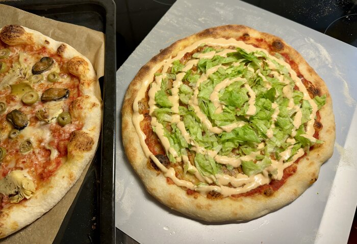 Två hembakade pizzor med olika toppings, sallad, sås, oliver och kronärtskocka, serveras på bakplåt och skärbräda.