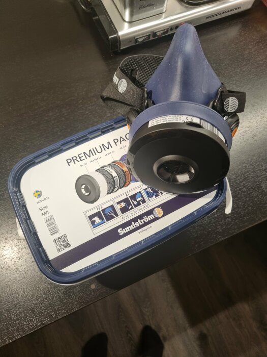 Blå andningsmask med filter ovanpå blå förpackning, på mörkt köksbänk bredvid kaffebryggare.