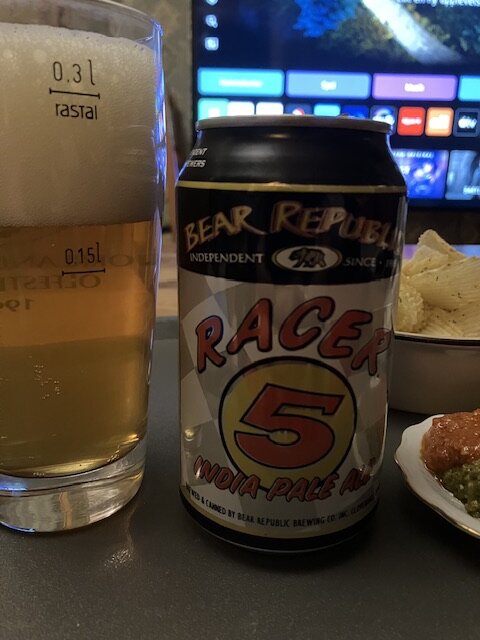 Öl i glas bredvid ölburk, "Racer 5 IPA", snacks, suddig bakgrund med datorskärmar.