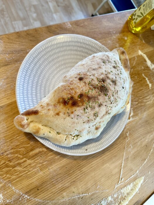 Nybakad calzone på präglat vitt tallrik, träskärbräda, mjöl, köksbord, olivolja i bakgrunden.