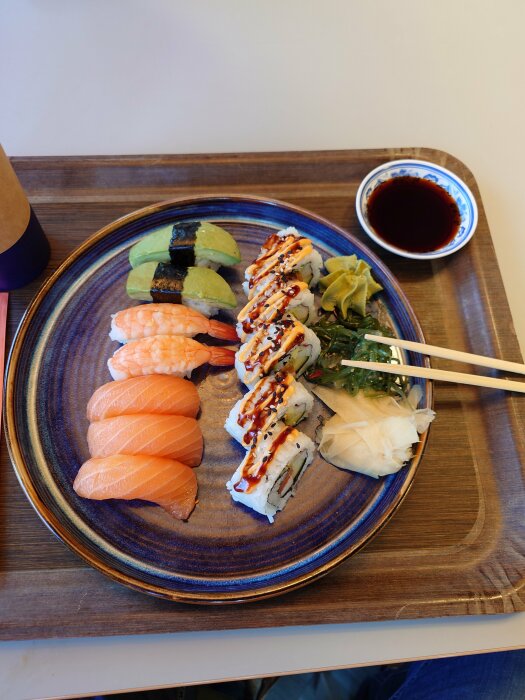 Sushitallrik med nigiri, maki, avokado, ingefära, wasabi, sojasås, ätpinnar på träunderlägg.