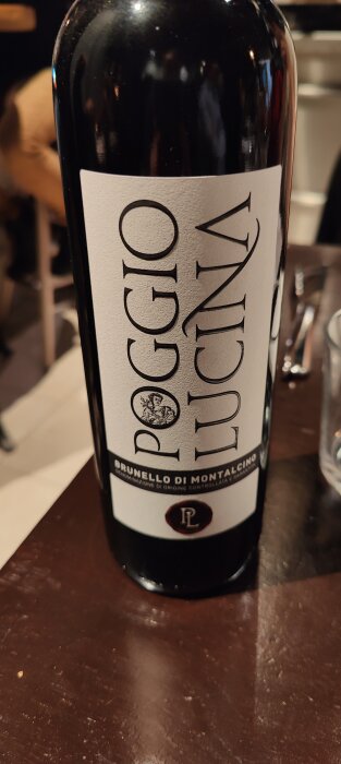 En vinflaska med etikett "POGGIO LUCINA Brunello di Montalcino", inget ansikte synligt, otydlig bakgrund.