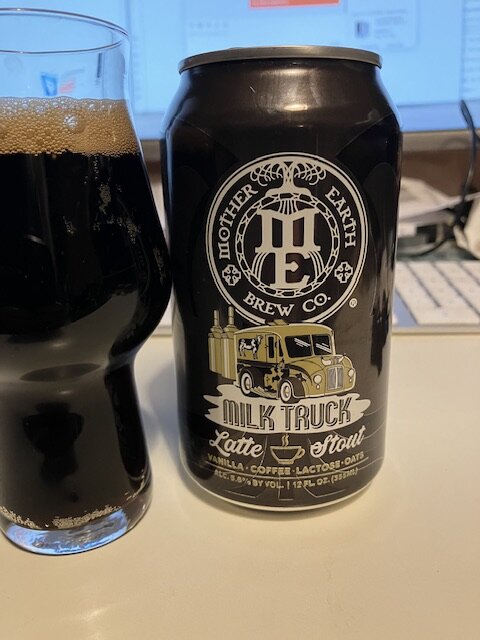 En burk Milk Truck Latte Stout bredvid ett glas med mörkt öl framför en datorskärm.