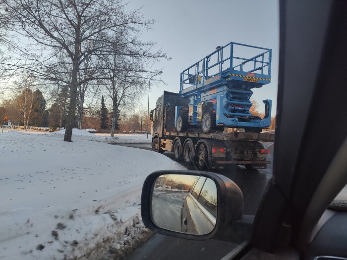 Lastbil transporterar skylift, tagen inifrån bil, vinter, greniga träd, snö, blå himmel.