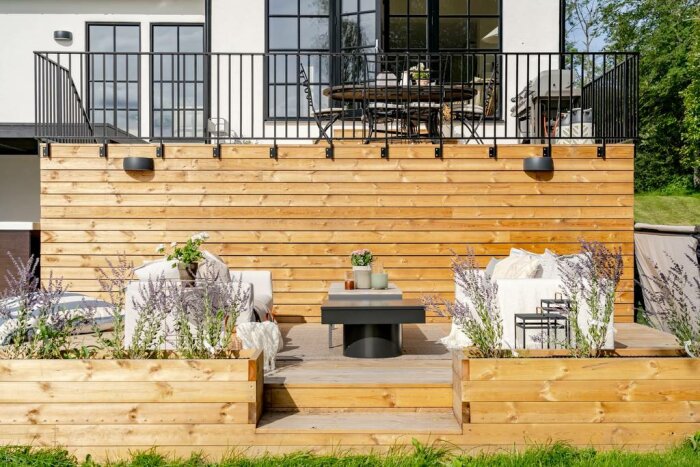 Modernt utomhusvardagsrum, träterrass, sittplatser, dekorativa plantor, balkong ovanför, stilrent design.