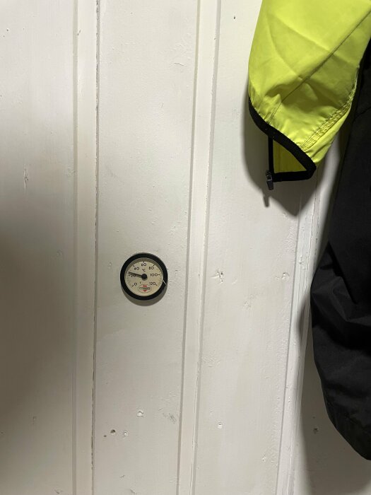 En vägg med en termometer och en del av en gul jacka.