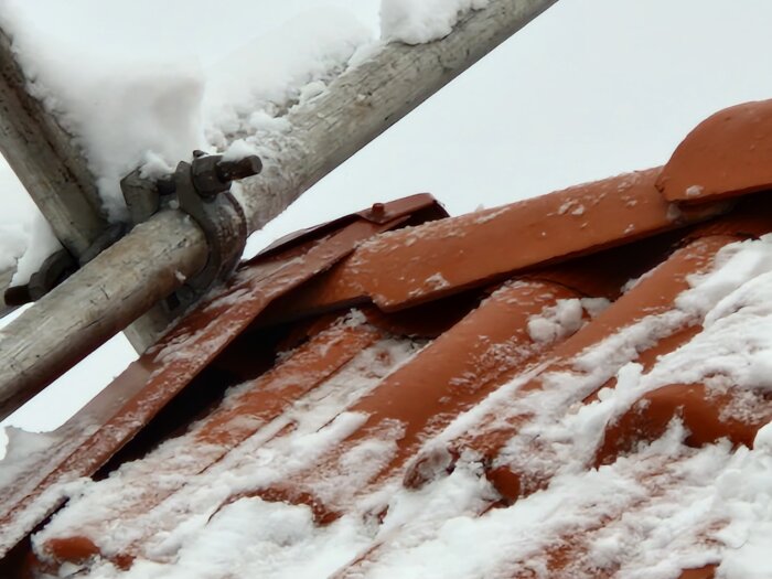 Snötäckt tak med röda tegelpannor och en metall-tvärstång. Kallt vinterväder.
