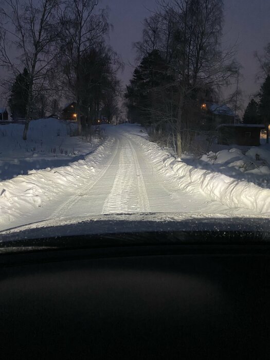 Vy från en bil vid snötäckt väg i skymningen med träd och hus i bakgrunden.