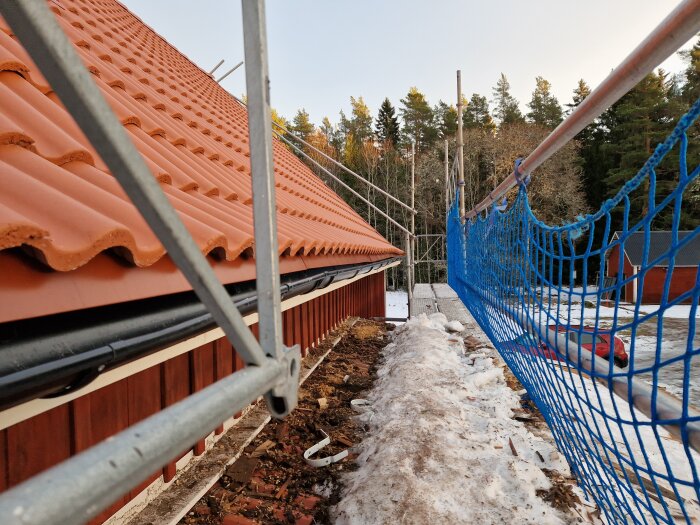 Byggarbetsplats med säkerhetsstaket, ställning, röd takpannor, smält snö, hus och träd i bakgrunden.