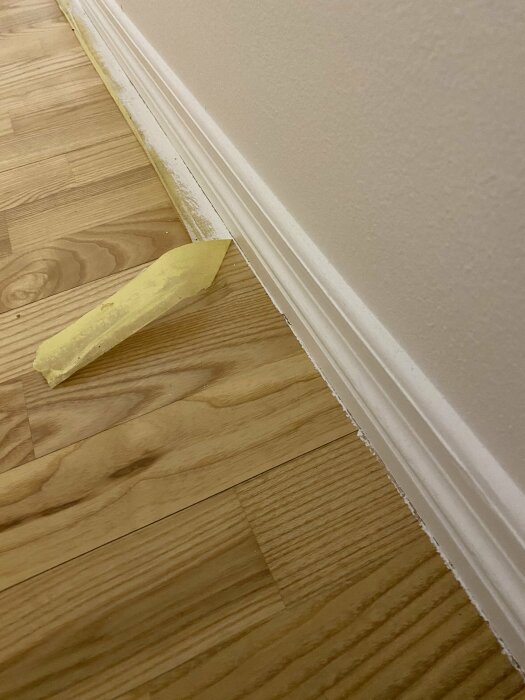 Golvhörn med träparkett, vit list, gul tejpremsa på golvet. Inomhus, närbild, byggnation eller renovering pågår eventuellt.