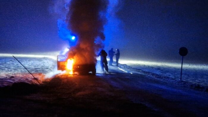 Bil brinner på snötäckt väg nattetid; personer iakttar; blåa ljus och rök; nödsituation; trafikskylt syns.