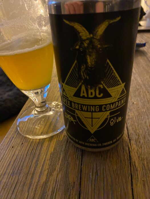 Ölglas med skum och en burk från Apex Brewing Company på ett bord.