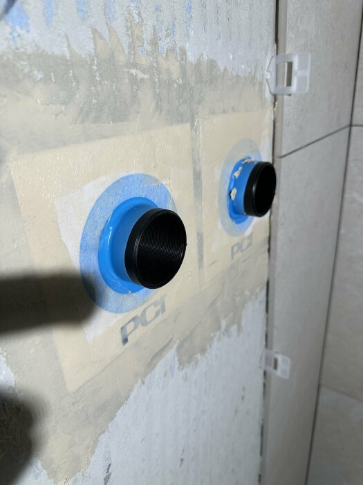 Två blå rörgenomföringar i en vägg under konstruktion, med kablar, kakel och isoleringsmaterial synligt.