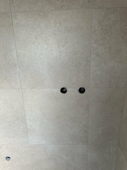 Vägg av ljusa stora kakelplattor, med två svarta inbyggda spotlights och avlopp i en dusch.