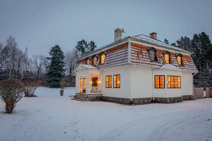 Ett stort hus i snölandskap vid skymning med upplysta fönster och vita pelare.