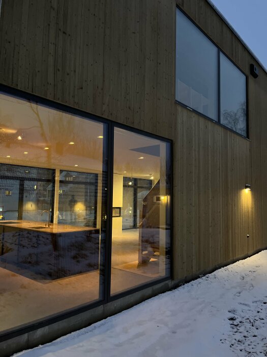 Modern byggnad med stora fönster, träfasad, belysning; snö täcker marken; skymningsljus.
