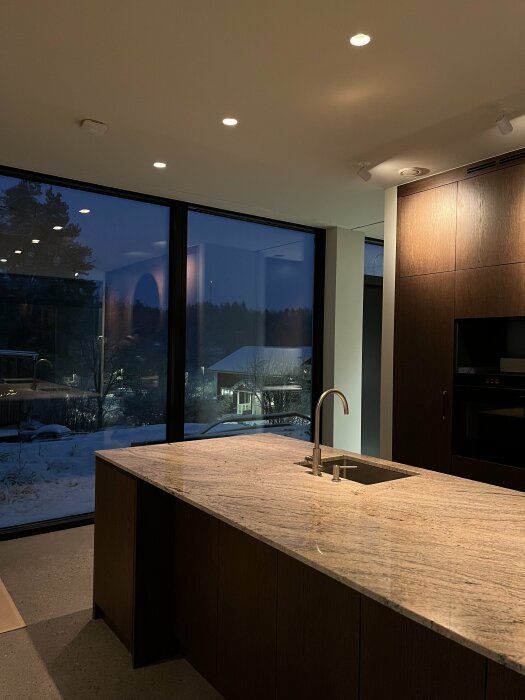 Modernt kök, stenbänk, inbyggda apparater, stora fönster, utsikt över vinterskymning, inomhusbelysning, reflektion, ordnat, rymligt.