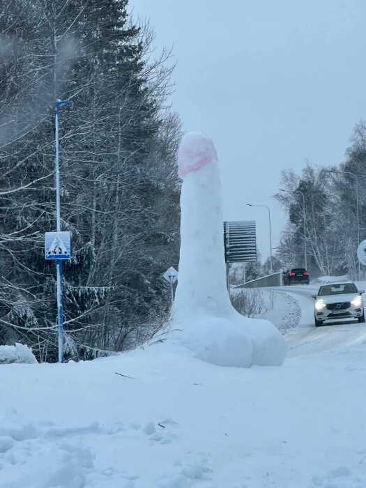 Snöskulptur med otydligt motiv, bilar på väg, vinterlandskap, busshållplats och skog i bakgrunden.