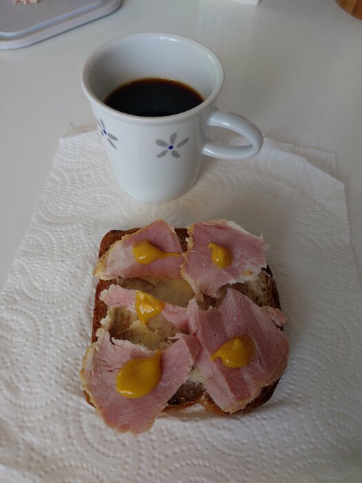 Kopp kaffe, macka med skinka och senap på servett.