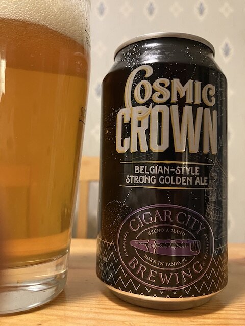 En ölburk märkt "Cosmic Crown", Belgian-style strong golden ale, bredvid ett glas med öl.