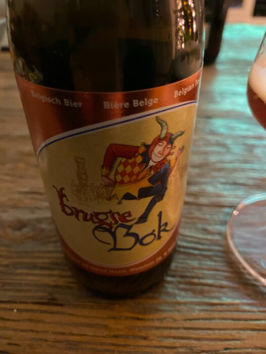 En ölflaska med etikett "Brugse Bok", illustrerad narrfigur, mot träbakgrund, text "Belgisch Bier". Glas syns suddigt.