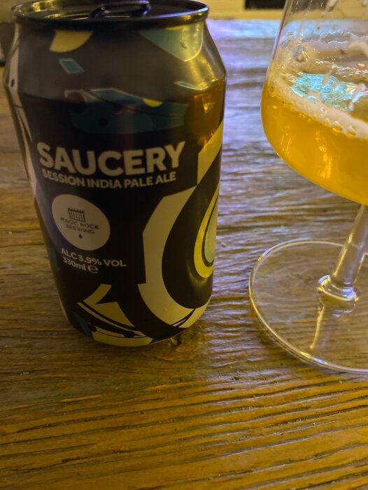 En ölburk märkt "SAUCERY Session India Pale Ale" bredvid ett glas med öl, på ett träbord.