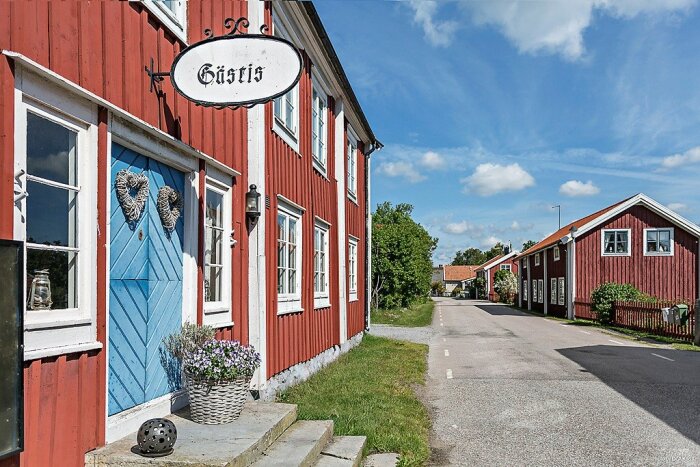 Röda trähus, blå dörr, skylt "Gästis", kullerstensväg, sommardag, gröna träd, traditionell svensk landsbygdsmiljö.