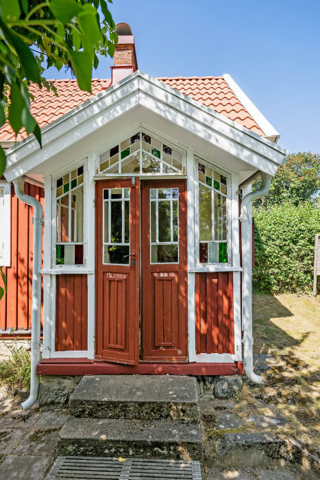 Charmigt litet rött hus med vita knutar, veranda och fönster, omgivet av grönska och blå himmel.