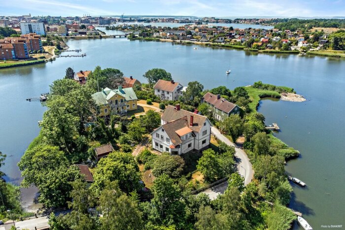 Flygbild på halvö med hus, träd, väg; omgiven av vatten och stadsbebyggelse i bakgrunden.