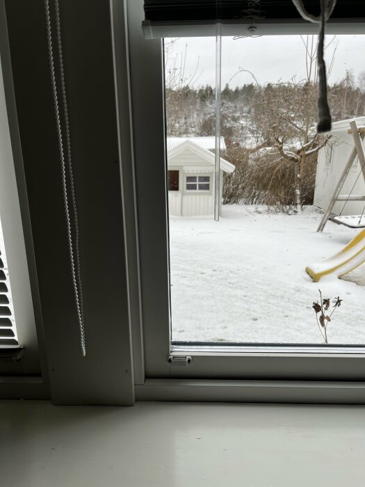 Vy genom öppet fönster mot snöklädd trädgård med byggnader och gunga. Grå himmel och vinterdag.