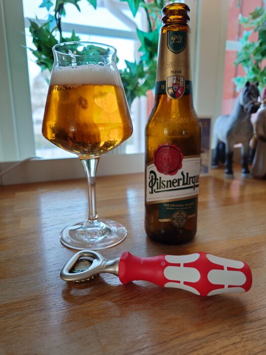 Ett glas öl, öppnad Pilsner Urquell-flaska och kapsylöppnare på ett träbord. Inomhus och dekorativa växter syns.