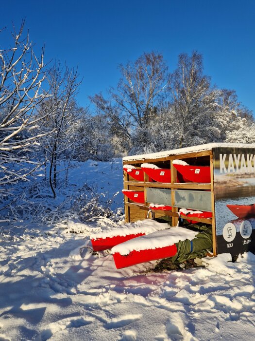 Röda kajaker lagrade i hylla, snötäckt mark, vintersceneri, träd med frost, klarblå himmel.