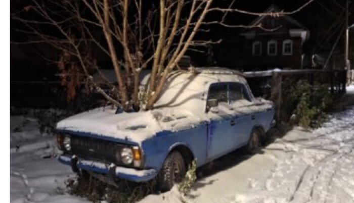 Gamla blå bilen täckt med snö, parkerad vid träbaumgivning, natt, med hus i bakgrunden.