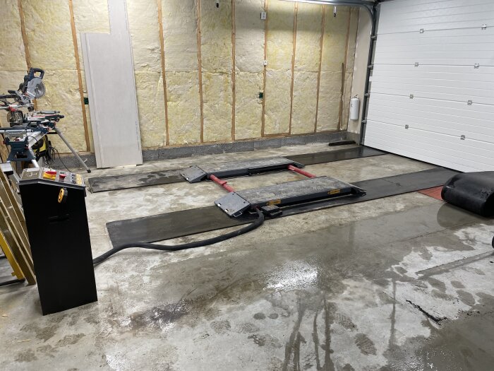 Garageverkstad med fordonslyft, isolering, verktyg, och vit garageport. Betonggolv med fettfläckar och kabeldragningar.