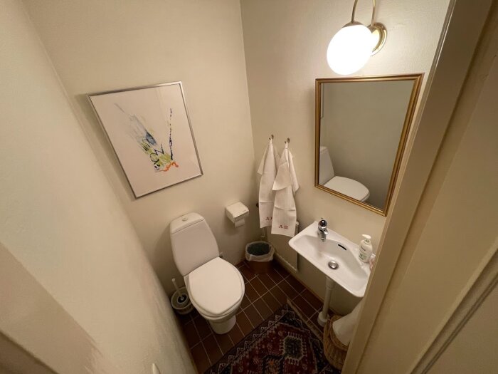 Toalett med handfat, spegel, konstverk, handdukar och belysning i ett litet rum.