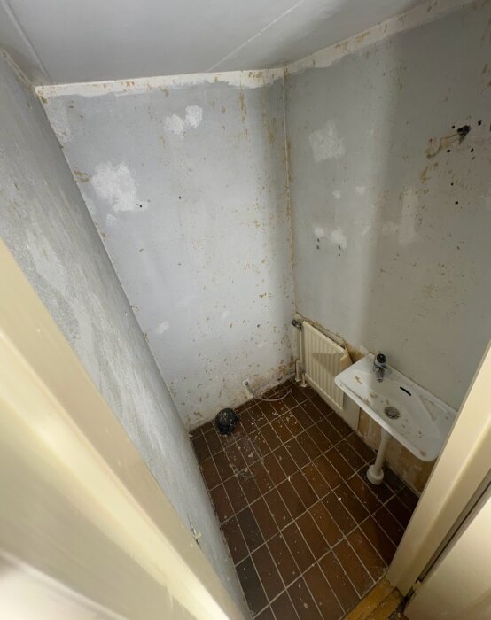 Slitet, smutsigt badrum med fläckiga väggar, handfat och brunt golv. Behov av renovering.