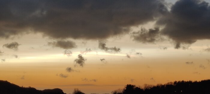 Solnedgång med mörka moln, ljusstrålar, silhuetter av kullar och träd, dramatisk himmel, orange och blå toner.