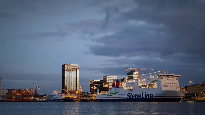 Hamn med Stena Line-färja, byggnader, mörka moln vid skymning, ljus från fönster och skepp.