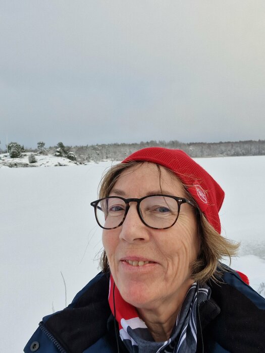 Kvinna i röd mössa ler, snöigt landskap i bakgrunden, natur, dagtid, utomhus, vinterkläder.