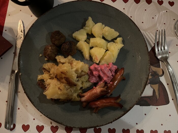 En tallrik med köttbullar, potatis, rödbetssallad, korv, potatisgratäng och sås, på hjärtmönstrad duk.